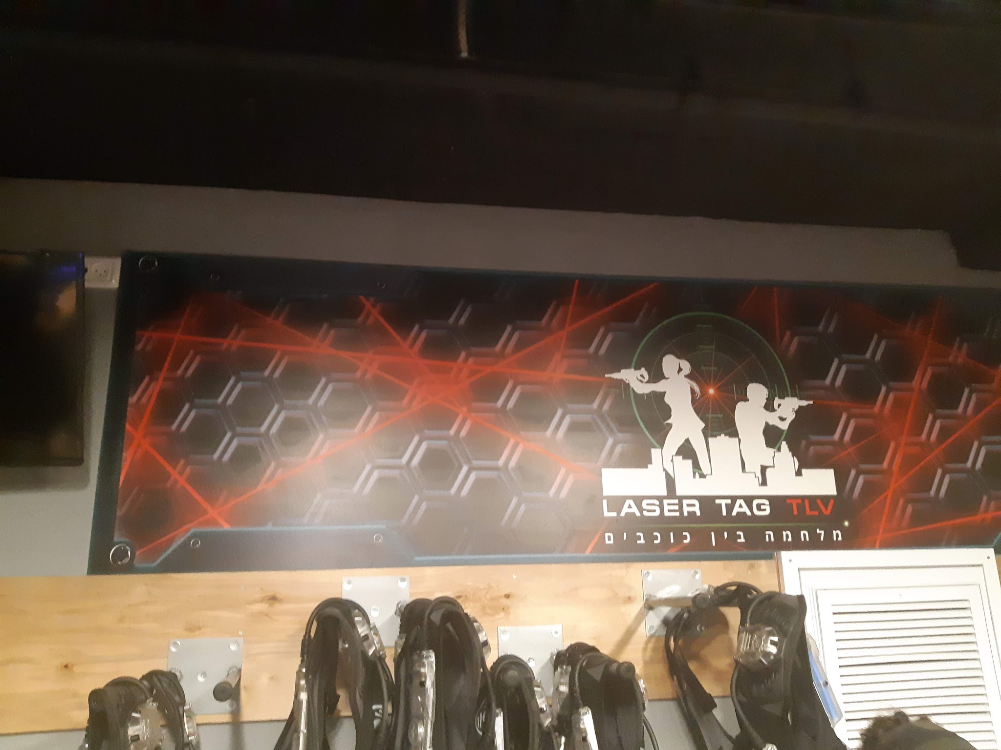שלט LASER TAG TLV עם הכיתוב מלחמה בין כוכבים, מעל מתלים של אפודים שחורים במתחם ורטיגו